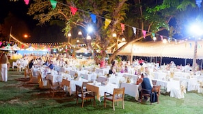 An outdoor event with multiple round tables covered with white tablecloths, surrounded by guests.