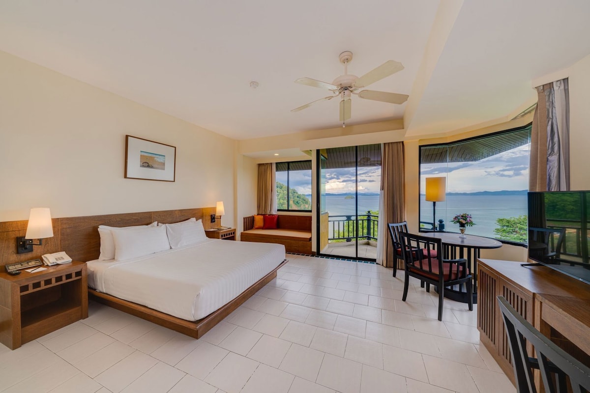 Habitación de hotel con cama, mesitas de noche, escritorio con silla, televisor y balcón con vistas.
