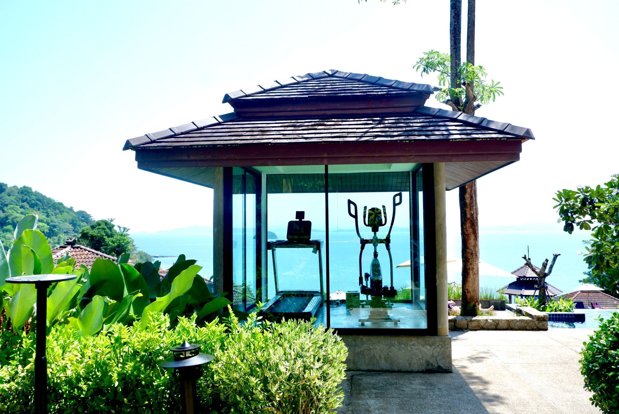 A glass-enclosed gazebo with a view of the sea, surrounded by lush greenery and a small pool.