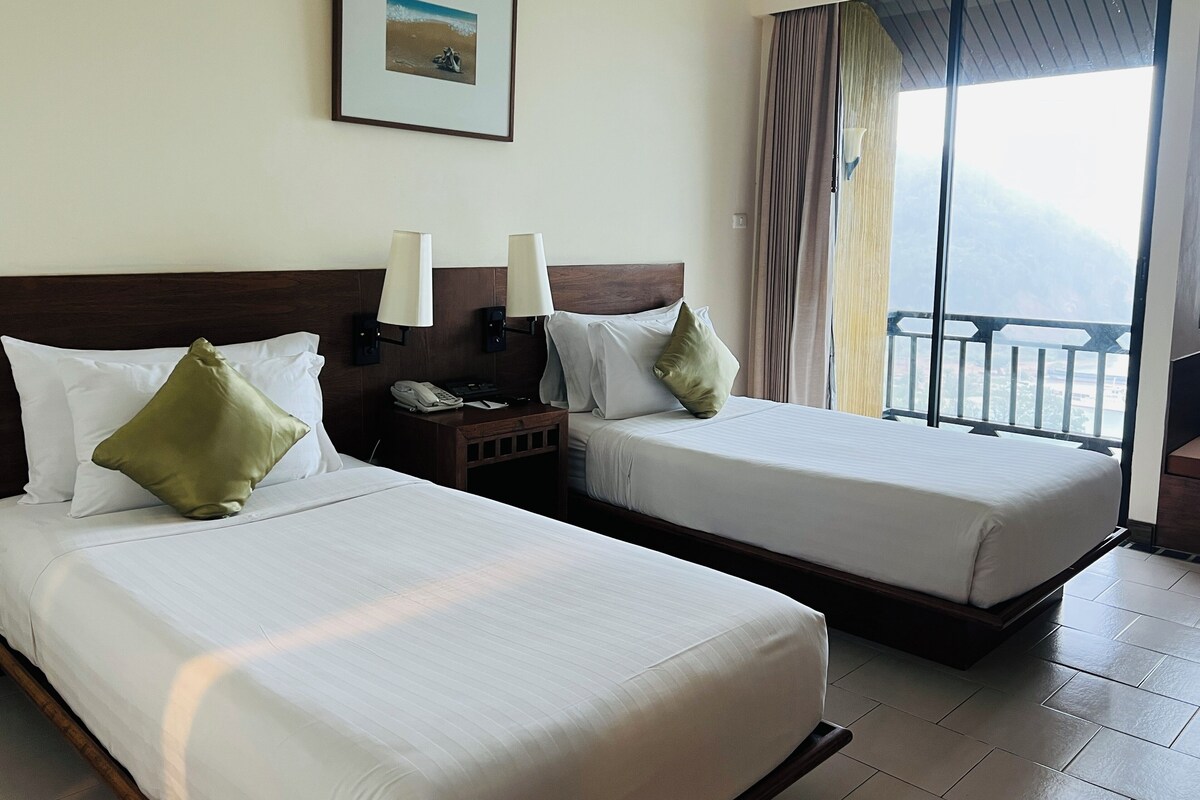 A hotel room with two beds, a nightstand with a phone, and a view of the sea through a balcony.