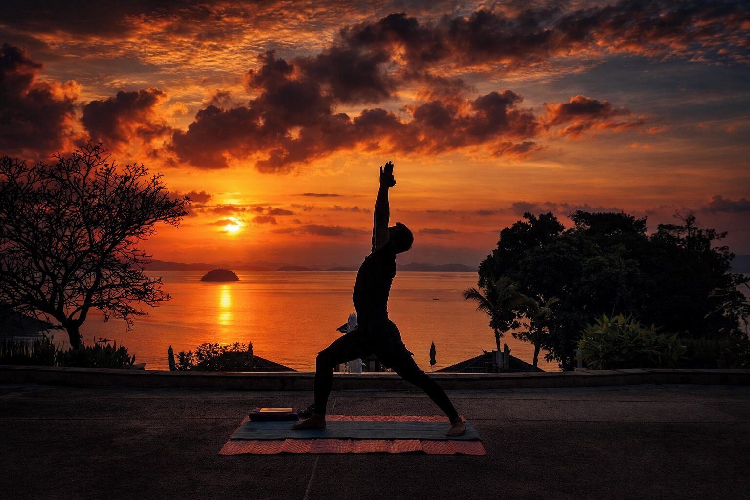 Uma pessoa praticando uma postura de yoga em um tapete, com vista para o pôr do sol e o oceano ao fundo.