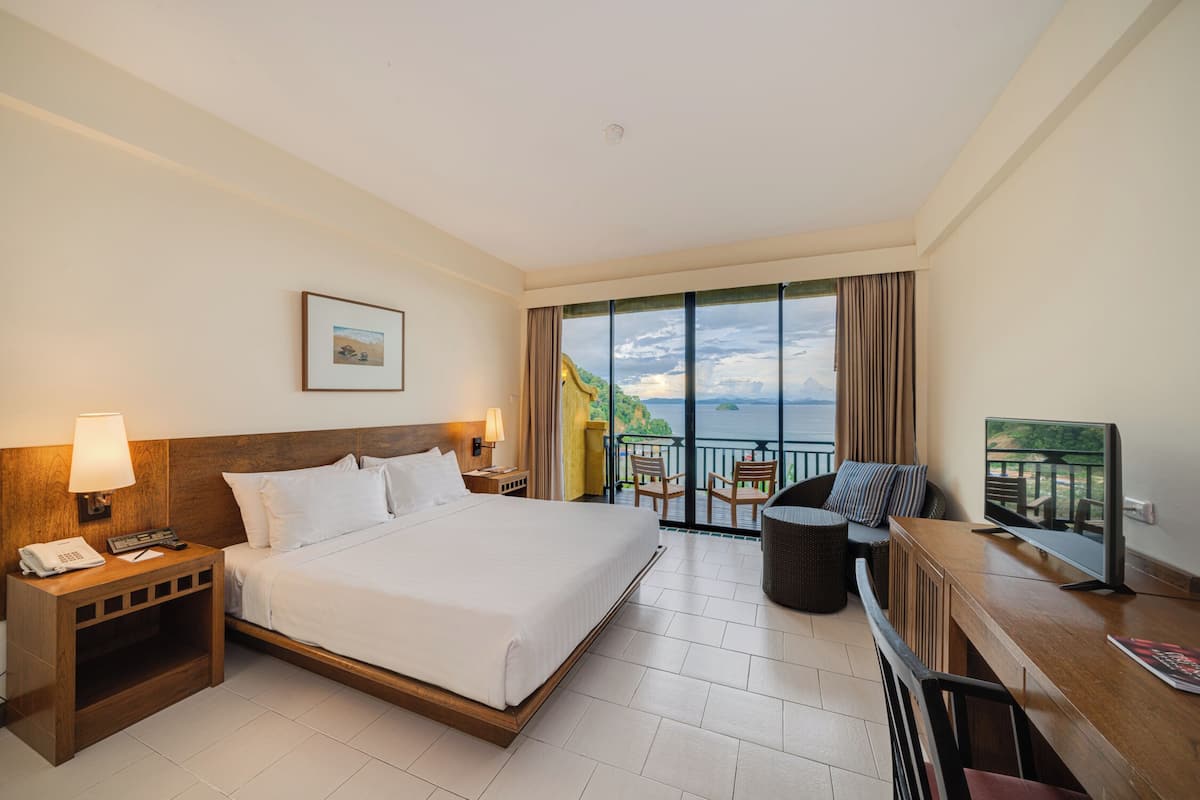 A hotel room with a large bed, a desk with a TV, a balcony with a view, and a small table with chairs.
