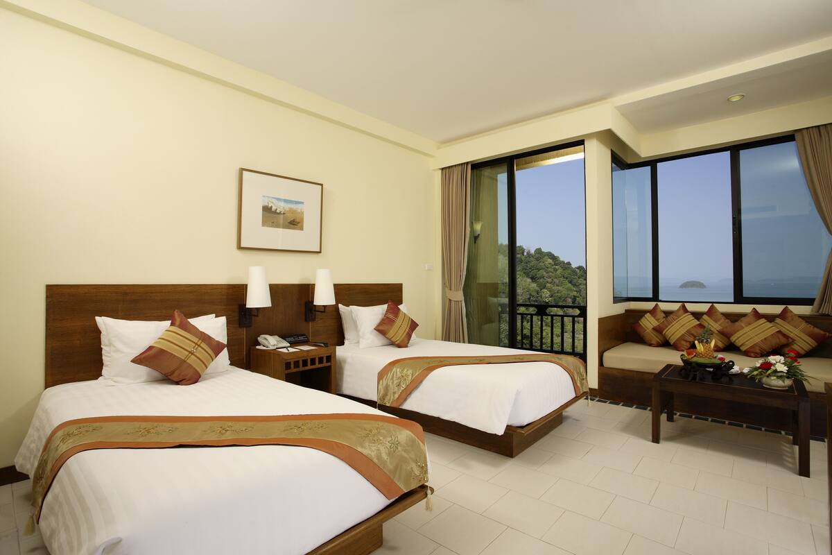 A hotel room with two beds, a sofa, a coffee table, and a large window offering a view.