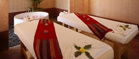 Two massage tables with towels and decorative pillows, a small sink, and a potted plant in a spa room.