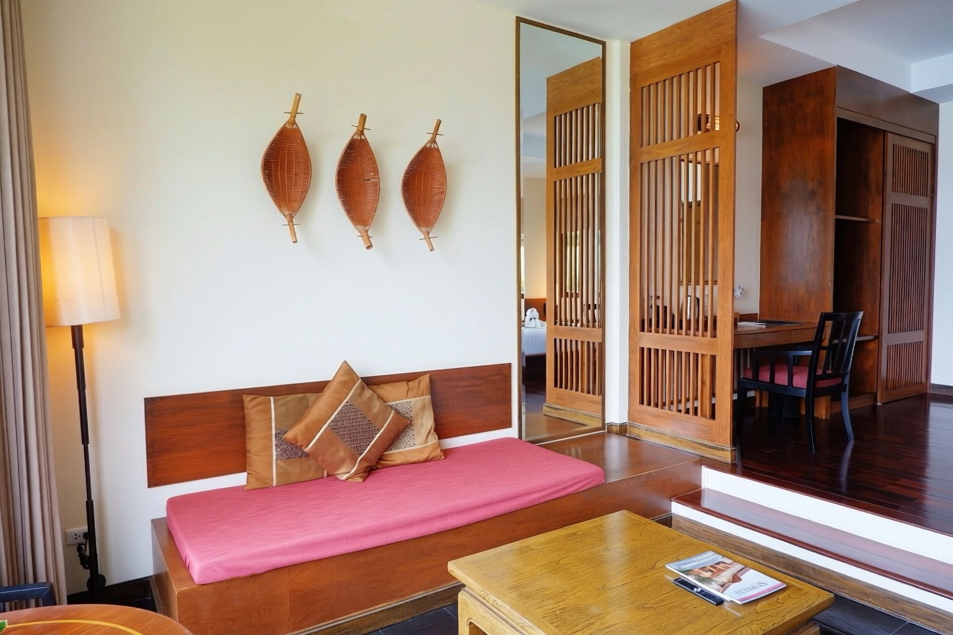 A hotel room with a wooden sofa, a small table, and a mirror reflecting a dining area.