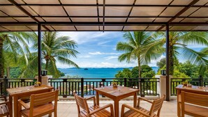 A covered outdoor seating area with wooden tables and chairs, overlooking a tropical landscape with palm trees and a view of the sea. - Supalai Scenic Bay Resort And Spa (Pa Klok)