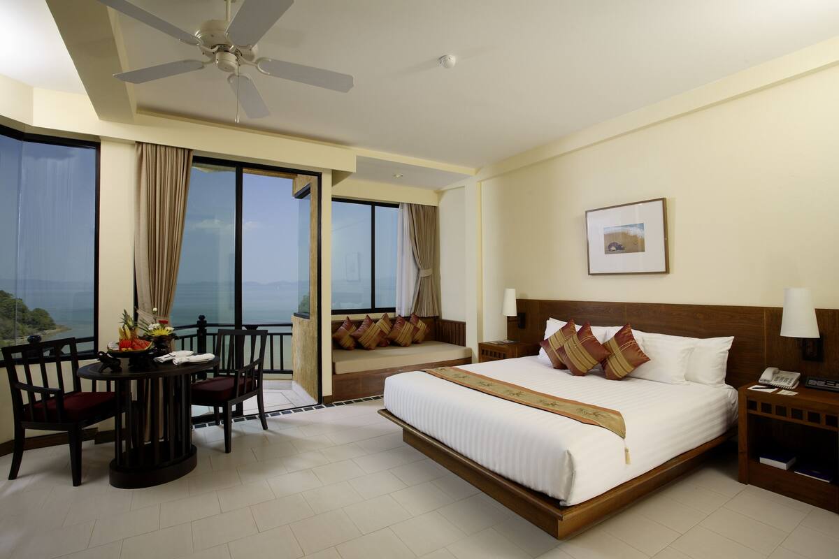 A hotel room with a large bed, a balcony with a view, a dining area with chairs, and a ceiling fan.