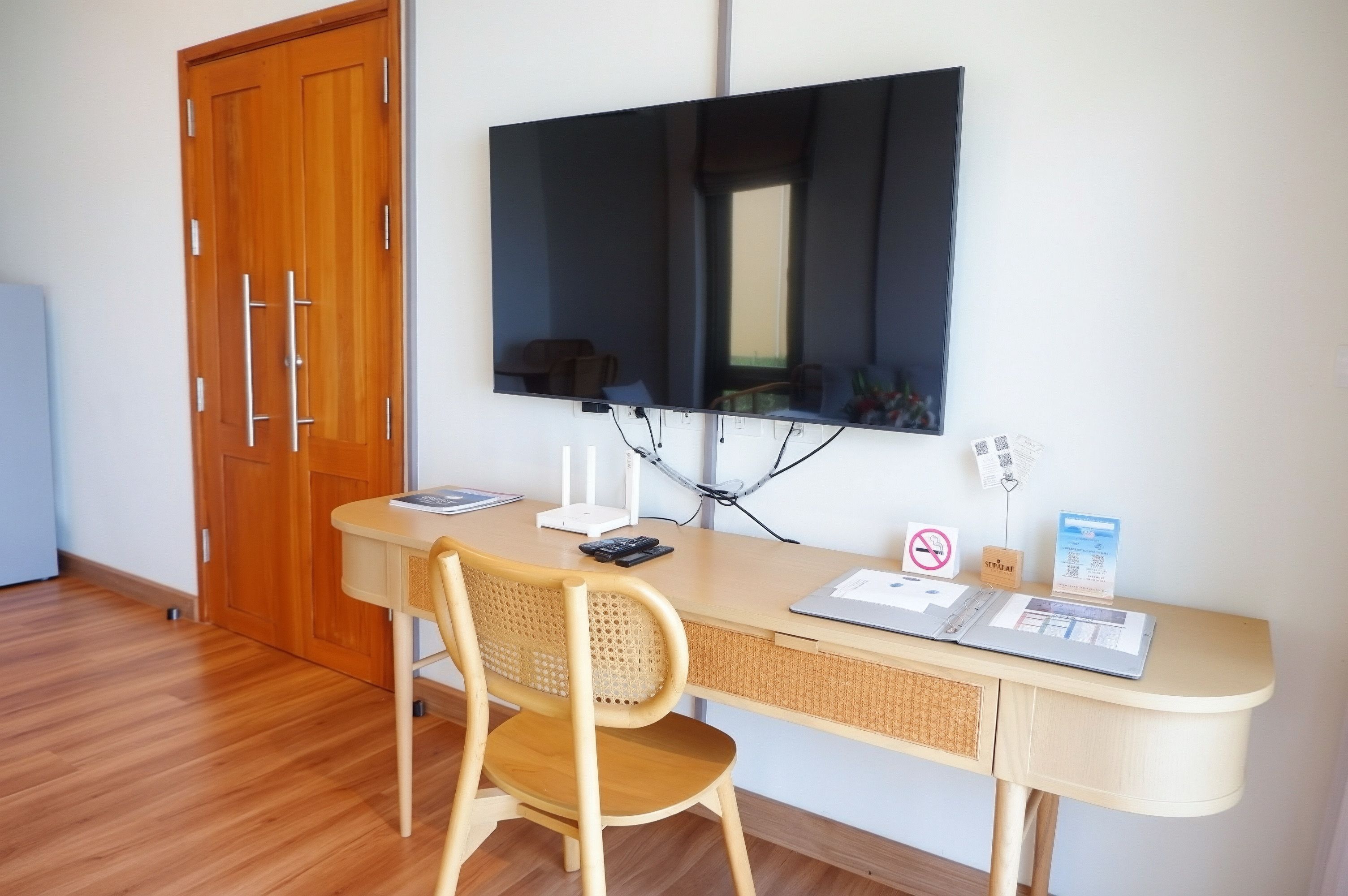 A room with a wooden desk, a chair, a flat-screen TV mounted on the wall, a door, and a window.