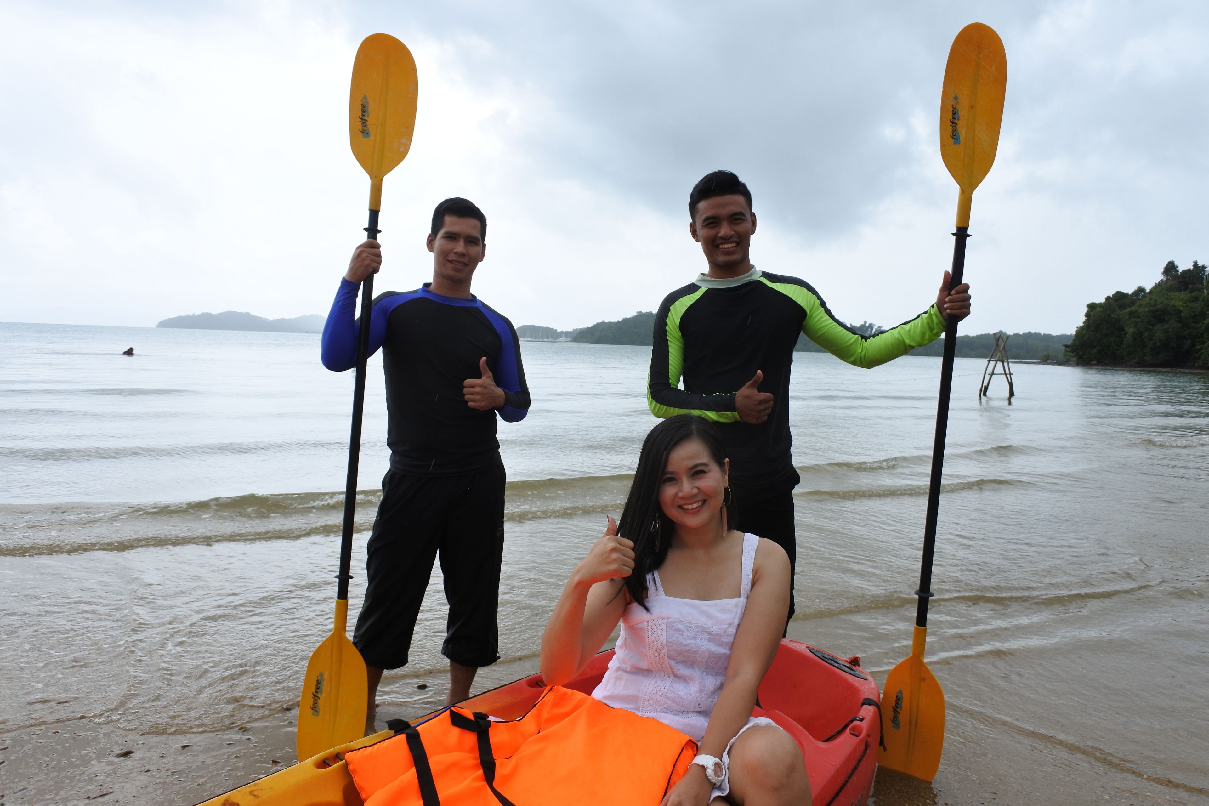 Three people holding paddles on a beach with a kayak and life jacket visible.