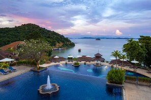 A resort pool with a fountain, lounge chairs, umbrellas, and a view of the sea and hills. - Supalai Scenic Bay Resort And Spa (Pa Klok)