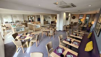 Un comedor de hotel moderno con diversas opciones de asientos, incluyendo mesas redondas y un sofá, además de una estación de buffet con exhibición de alimentos.