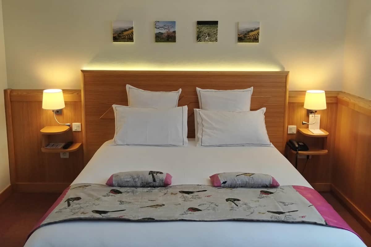 Ein Hotelzimmer mit einem hölzernen Kopfteil, einem großen Bett mit weißen Leinentüchern, zwei Nachttischen mit Lampen und drei gerahmten Bildern an der Wand.
