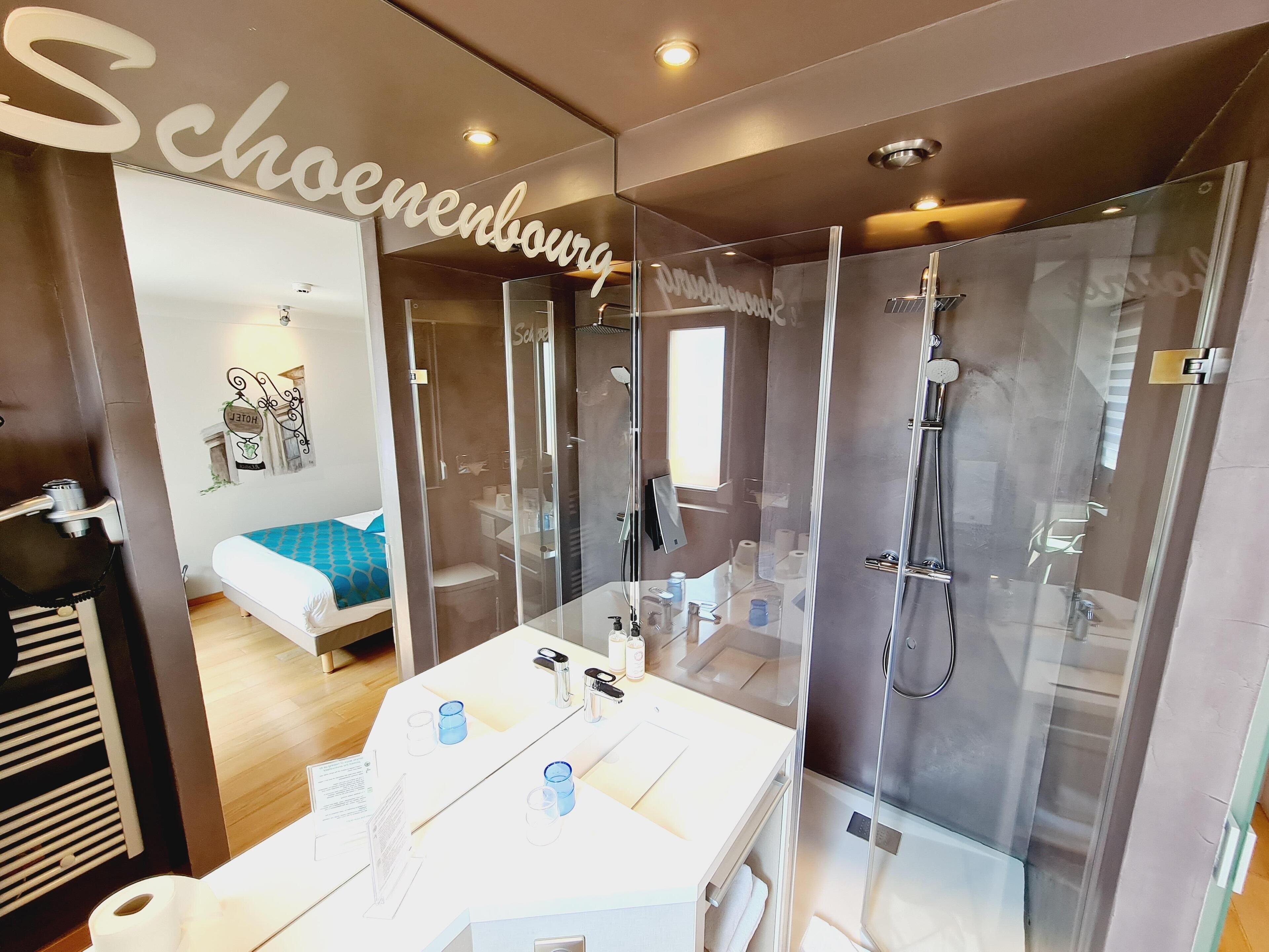 Un baño moderno con cabina de ducha de vidrio, un lavamanos blanco con artículos de aseo y una cama con colcha azul estampada.