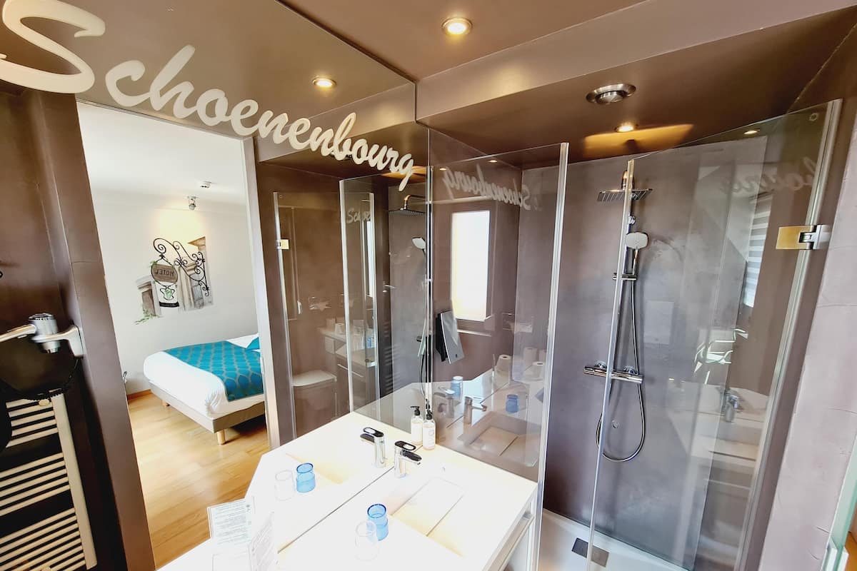 Ett modernt badrum med en duschkabin i glas, en vit tvättställskommod med hygienartiklar och en säng med ett blått, mönstrat överkast.