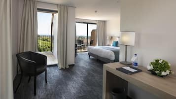 Habitación de hotel con cama, escritorio, silla, balcón y vista a zonas verdes.