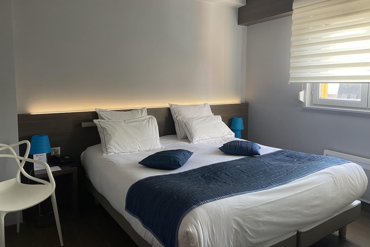 Ein Hotelzimmer mit Bett, Nachttischen, einem Sessel und einem Fenster mit Jalousien.