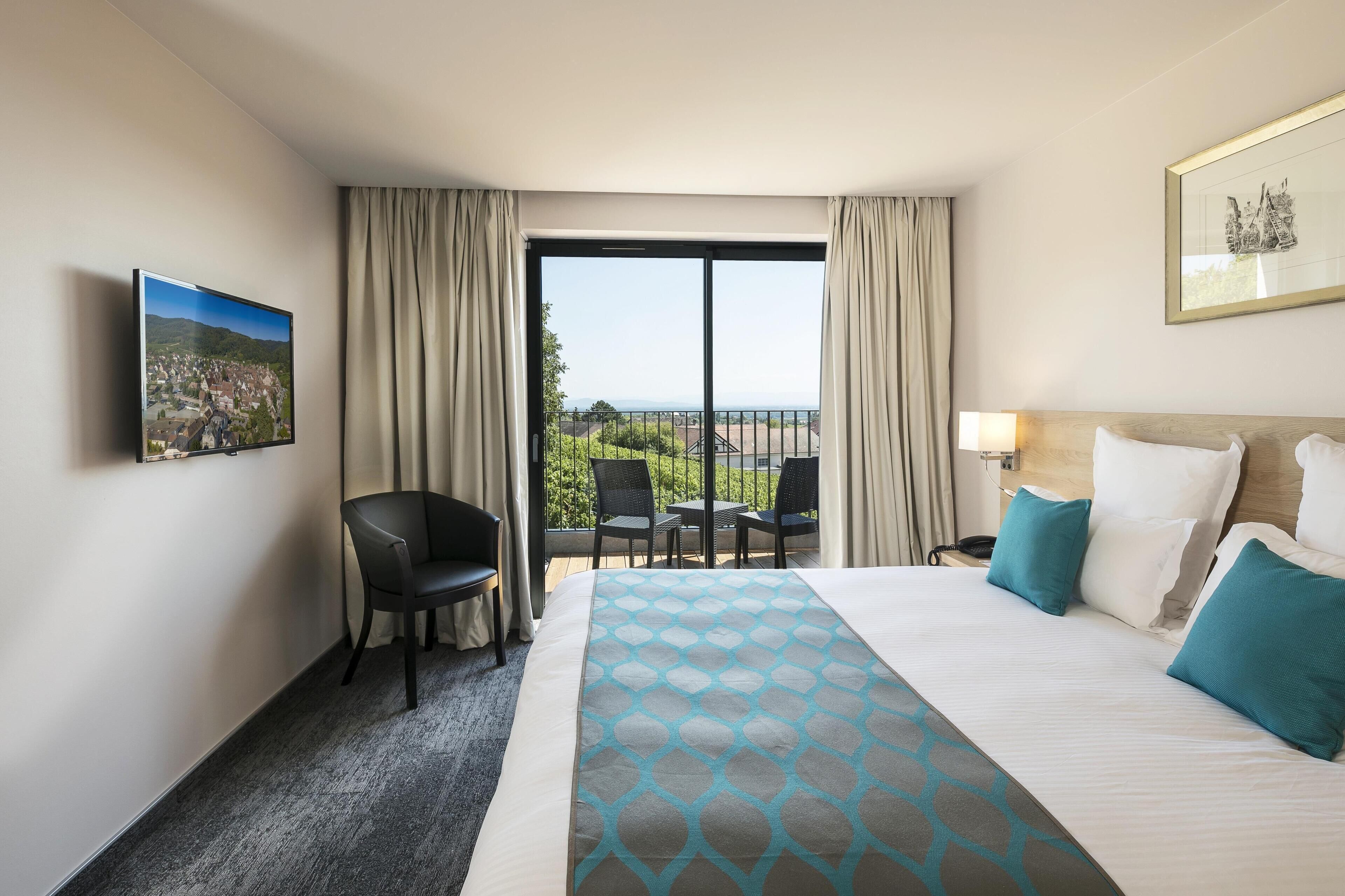 A hotel room with a bed, a desk, a chair, a TV, and a balcony with a view.