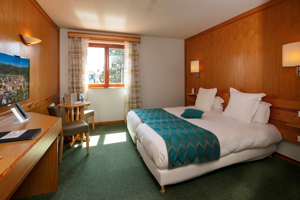 Ein Hotelzimmer mit Bett, Schreibtisch, Stuhl, einem Fenster mit Vorhängen und einem Fernseher an der Wand.