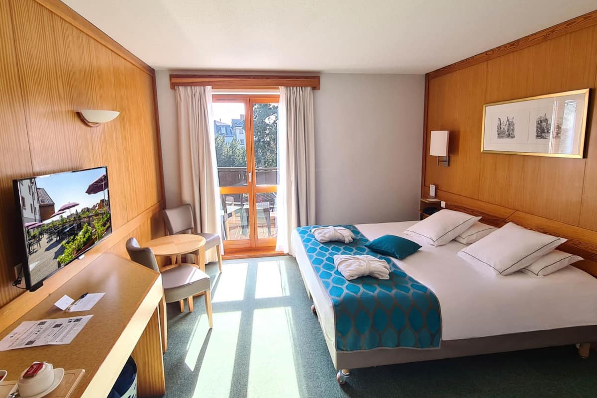 Ein Hotelzimmer mit Bett, Schreibtisch, Stuhl, Fernseher und einem Balkon mit Vorhängen.