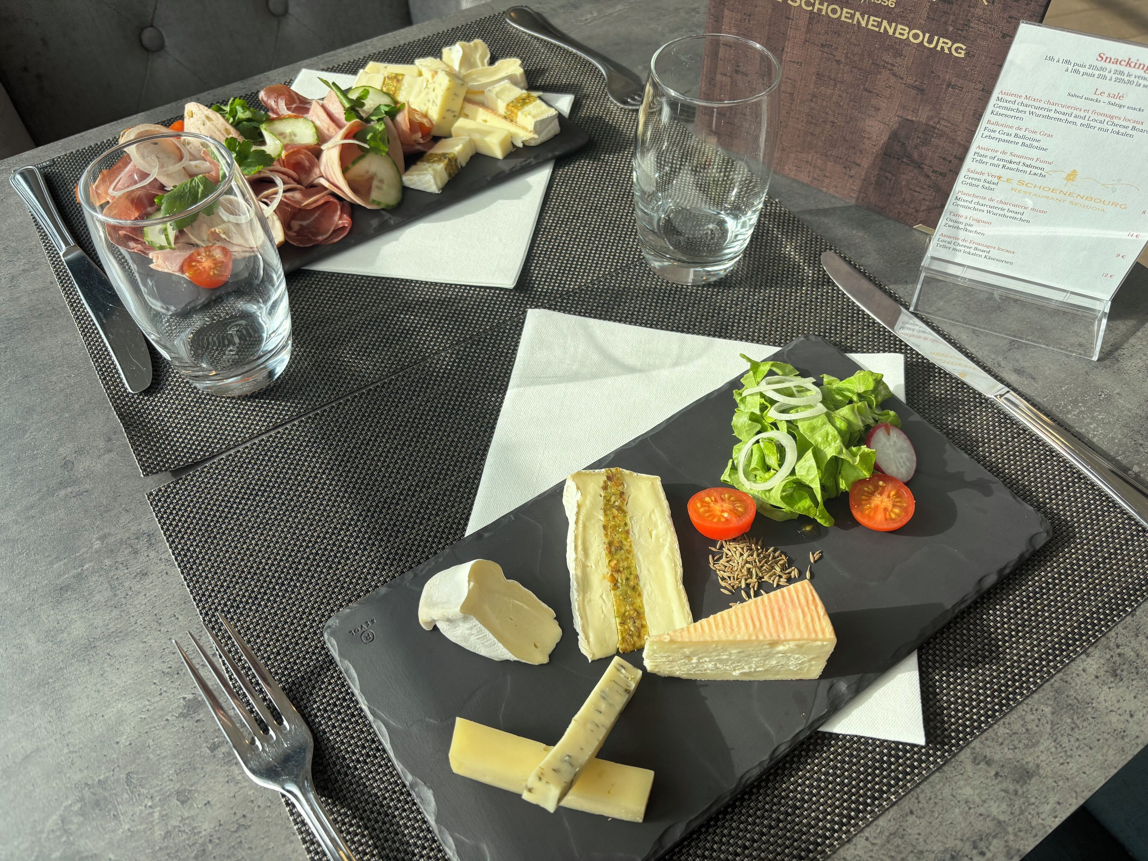 Ein Tisch mit einer Auswahl an Käse, Wurstwaren und Gemüse, dazu ein Glas Wasser und eine Speisekarte.