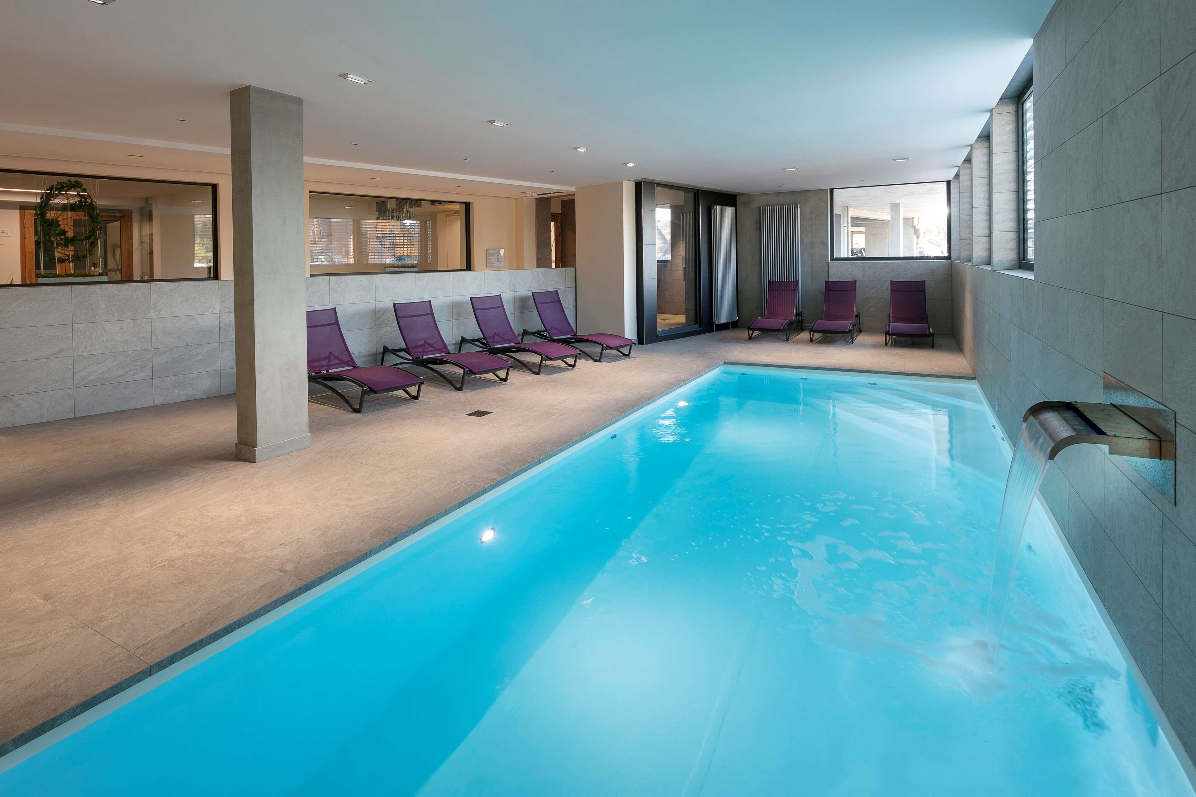 A modern indoor swimming pool with clear blue water, surrounded by lounge chairs and a large window providing natural light.