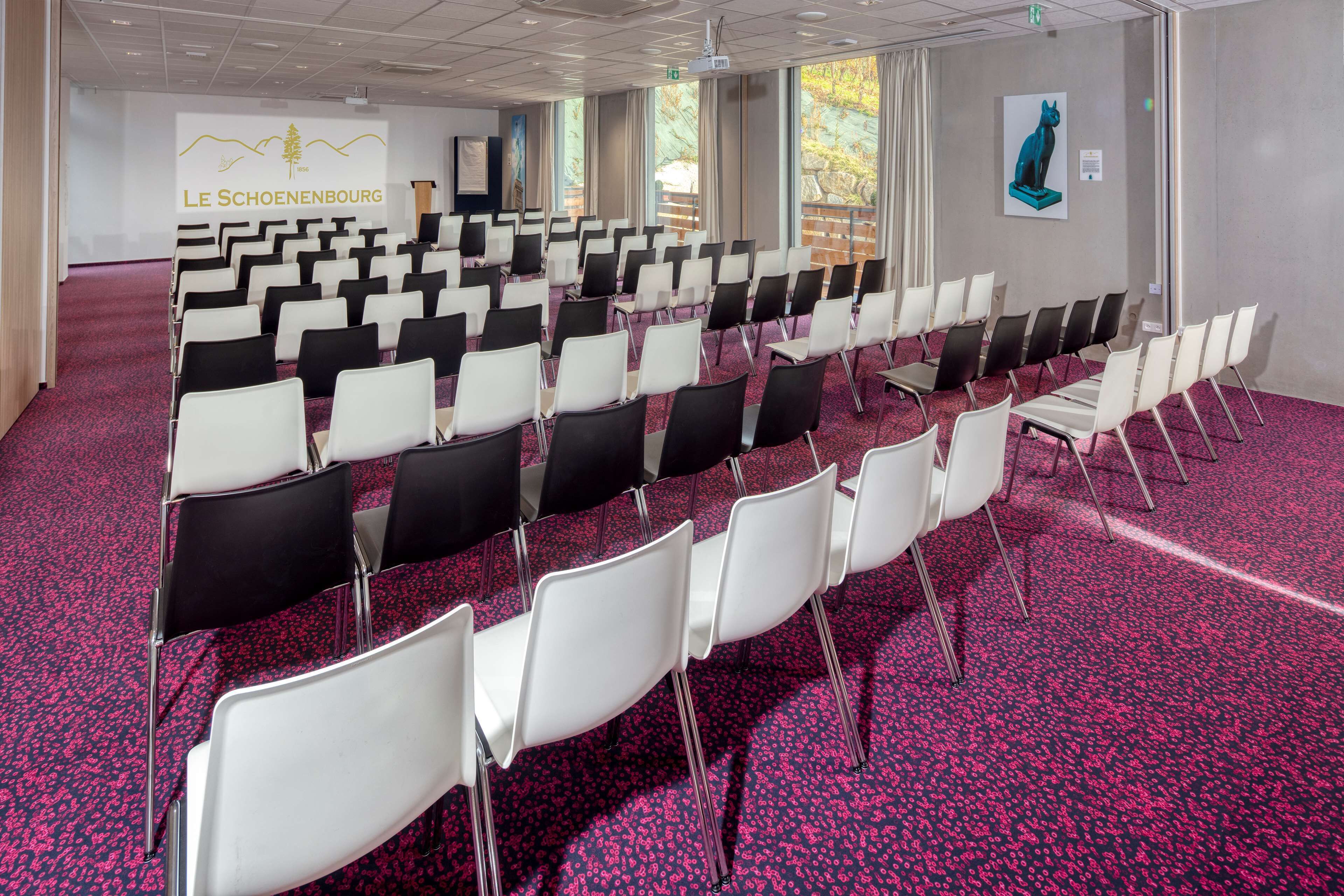 Un salón de conferencias con filas de sillas, piso alfombrado y un amplio ventanal.