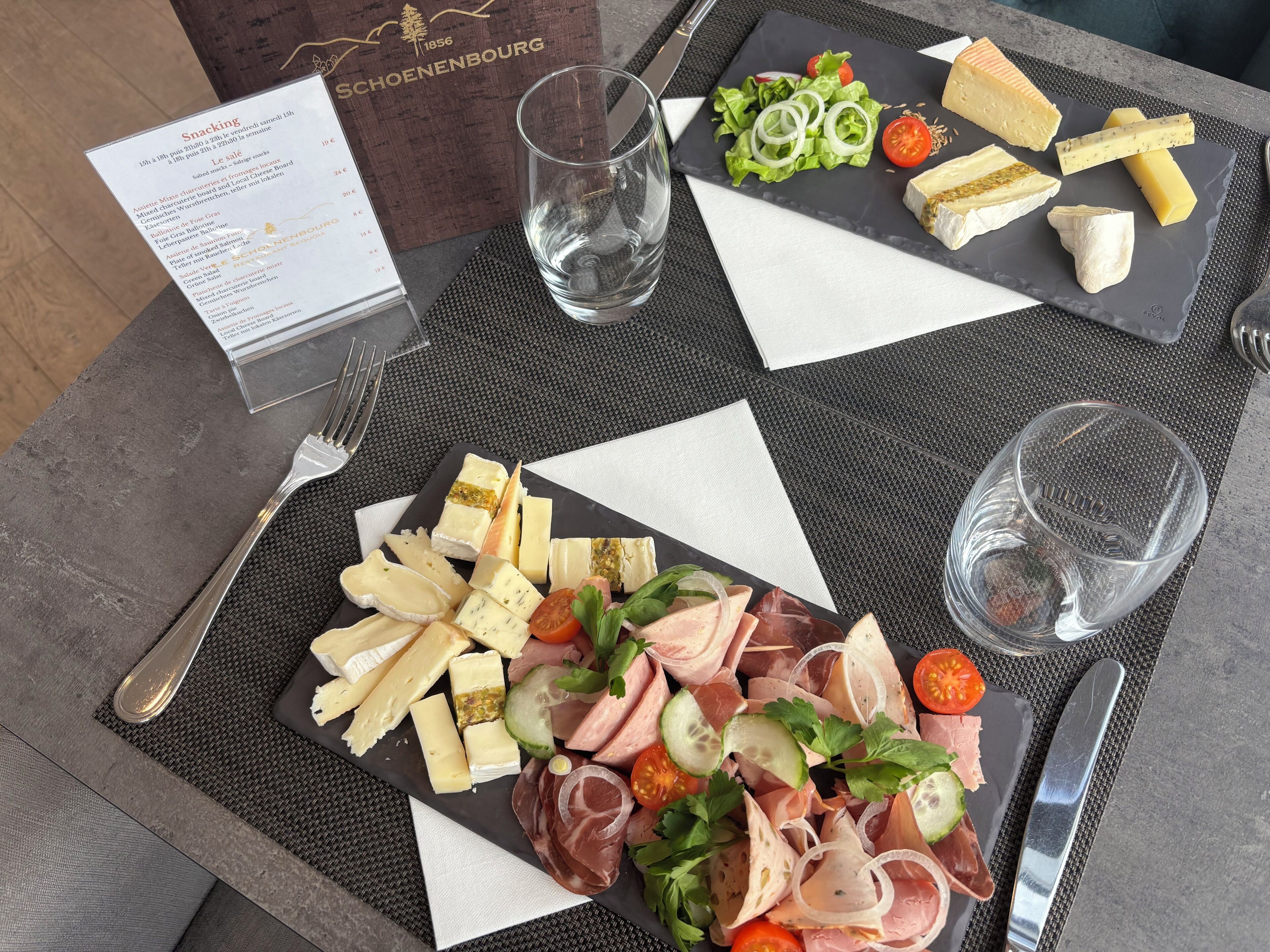 Une table garnie d’une variété de fromages, de charcuteries et d’une salade, accompagnée d’un menu et de verres.