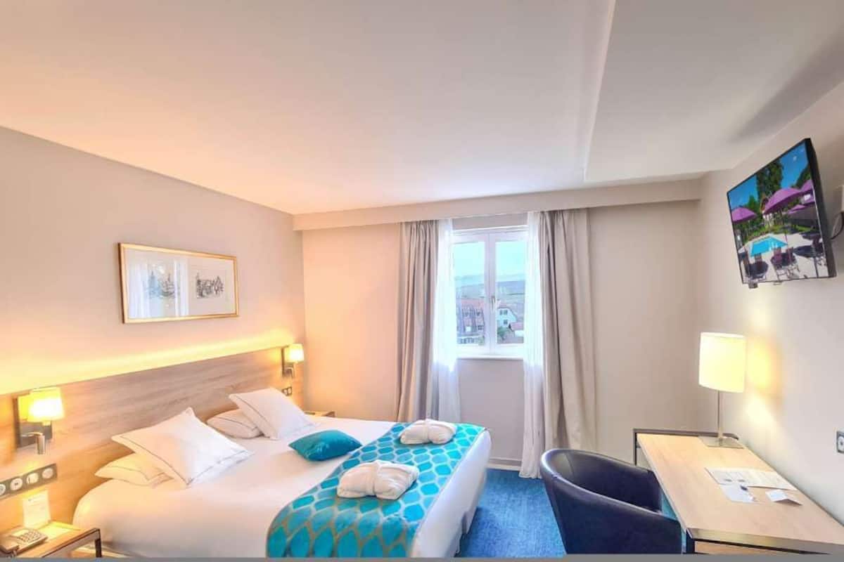 Ein Hotelzimmer mit Bett, Schreibtisch, Stuhl, einem Fenster mit Vorhängen und einem Bild an der Wand.