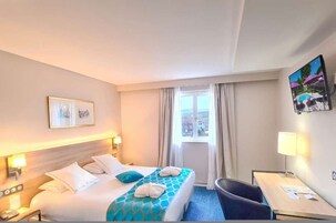 Habitación de hotel con cama, escritorio, silla, ventana con cortinas y un cuadro en la pared.
