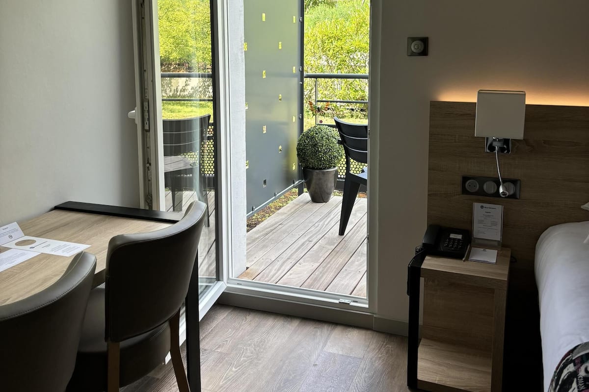 Ein Hotelzimmer mit Holzboden, einem Schreibtisch mit Papieren, einem Sessel, einem Bett, einem Nachttisch mit Lampe und Blick auf einen Balkon mit Tisch und Stühlen.