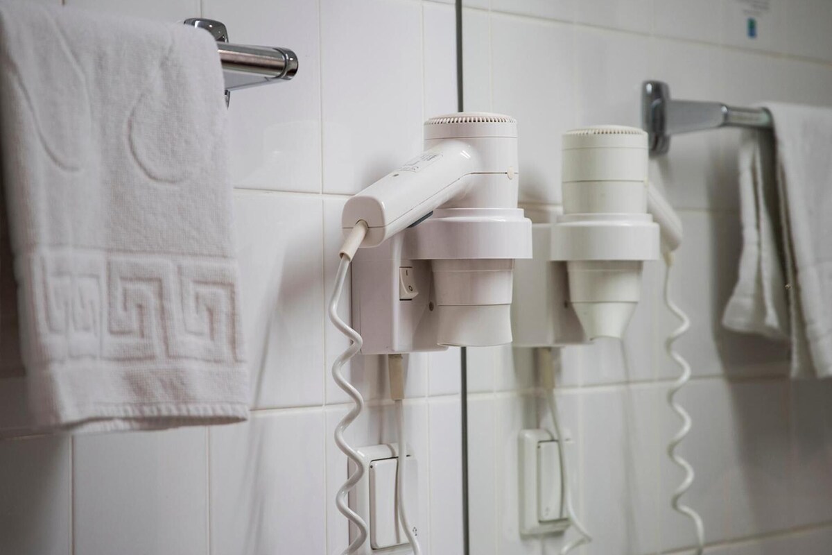 A bathroom with white tiles, a towel rack with a white towel, and two hair dryers mounted on the wall.