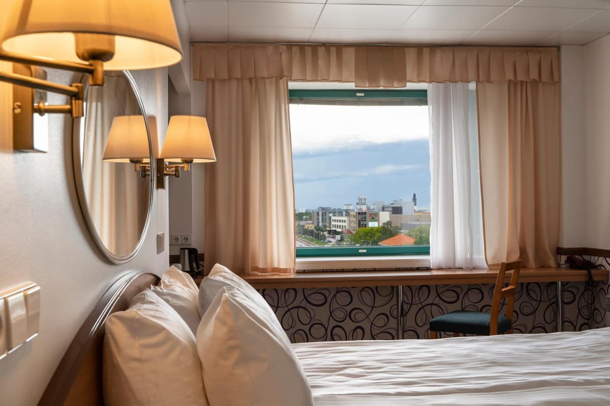 Una habitación de hotel con cama, una silla, un escritorio, una lámpara y una ventana con cortinas.