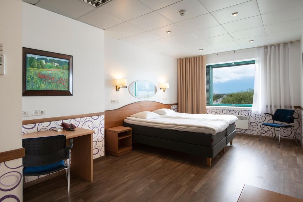 Habitación de hotel con cama, mesita de noche, silla, escritorio y ventana con vistas.