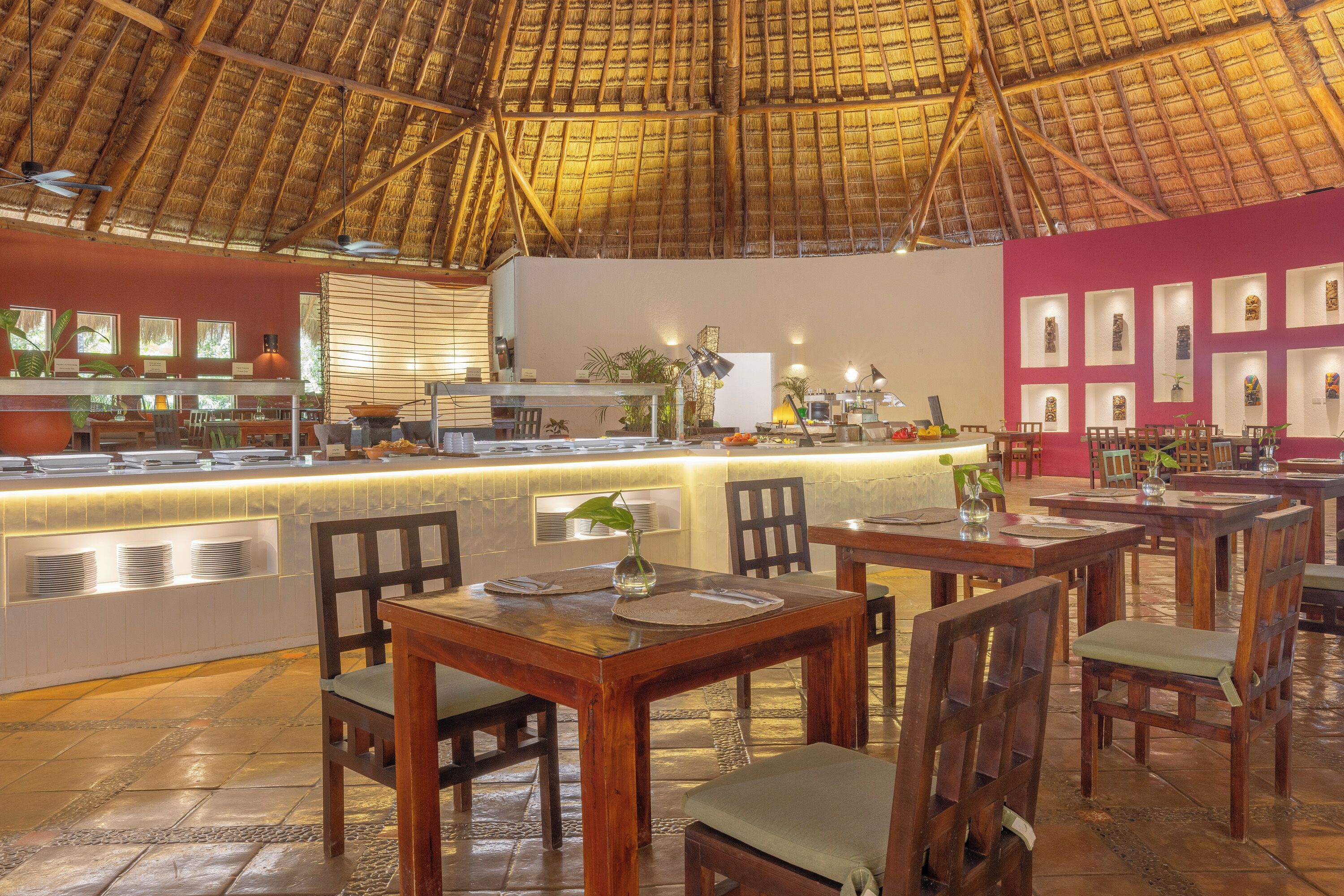 A restaurant with a thatched roof, wooden tables and chairs, a counter with a display of plates, and framed pictures on the wall.