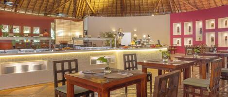 A restaurant with a thatched roof, wooden tables and chairs, a counter with a display of plates, and framed pictures on the wall.