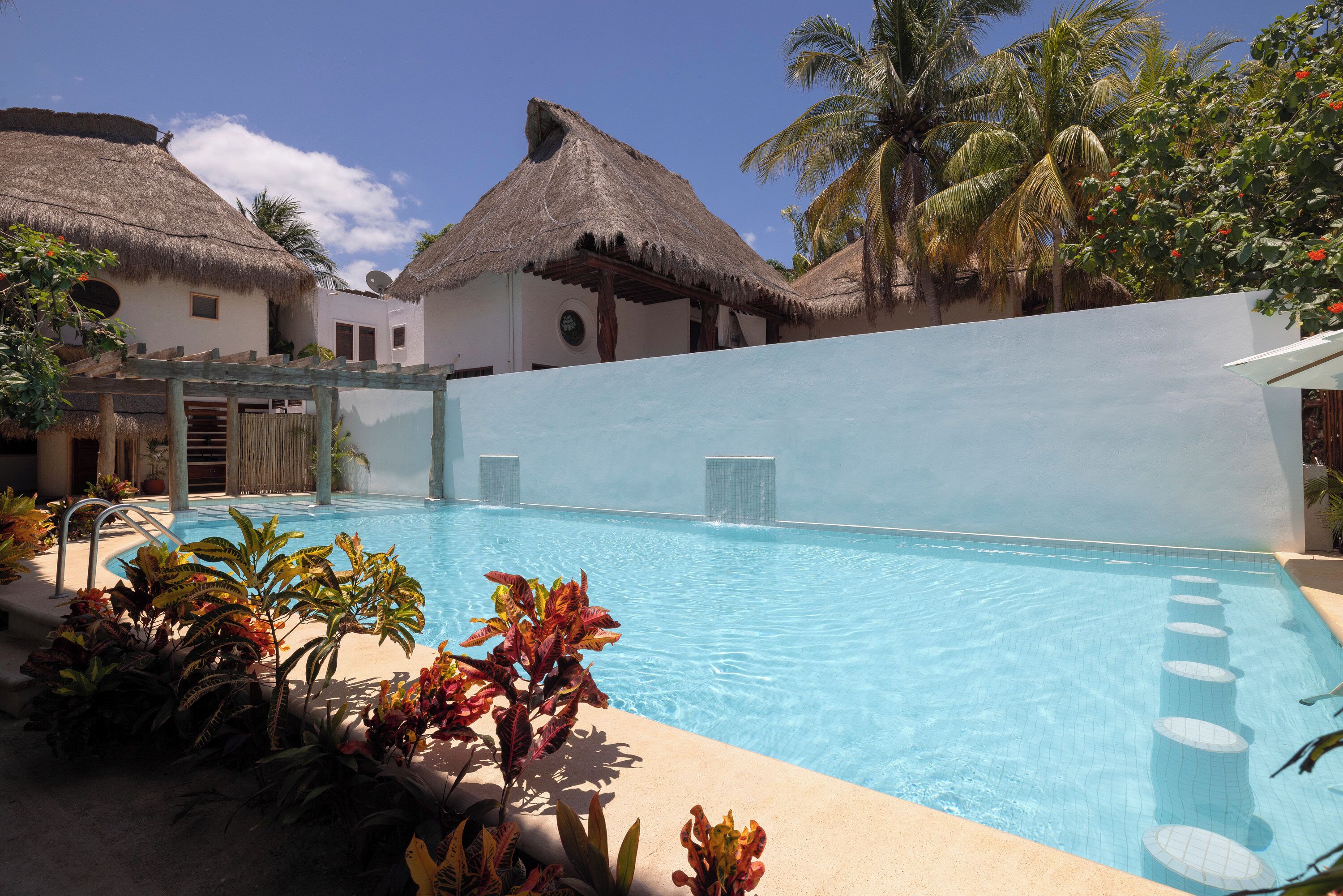 Une piscine dotée d’une structure surmontée d’un toit de chaume, de palmiers et de plantes tropicales.