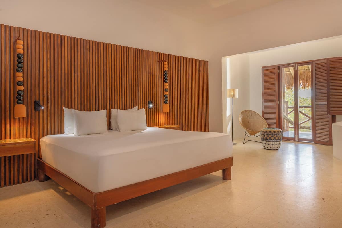Ein Schlafzimmer mit einer Wand aus Holzlatten, einem Bett mit weißen Leinentüchern, einem Nachttisch, einer Lampe und einem Korbstuhl.