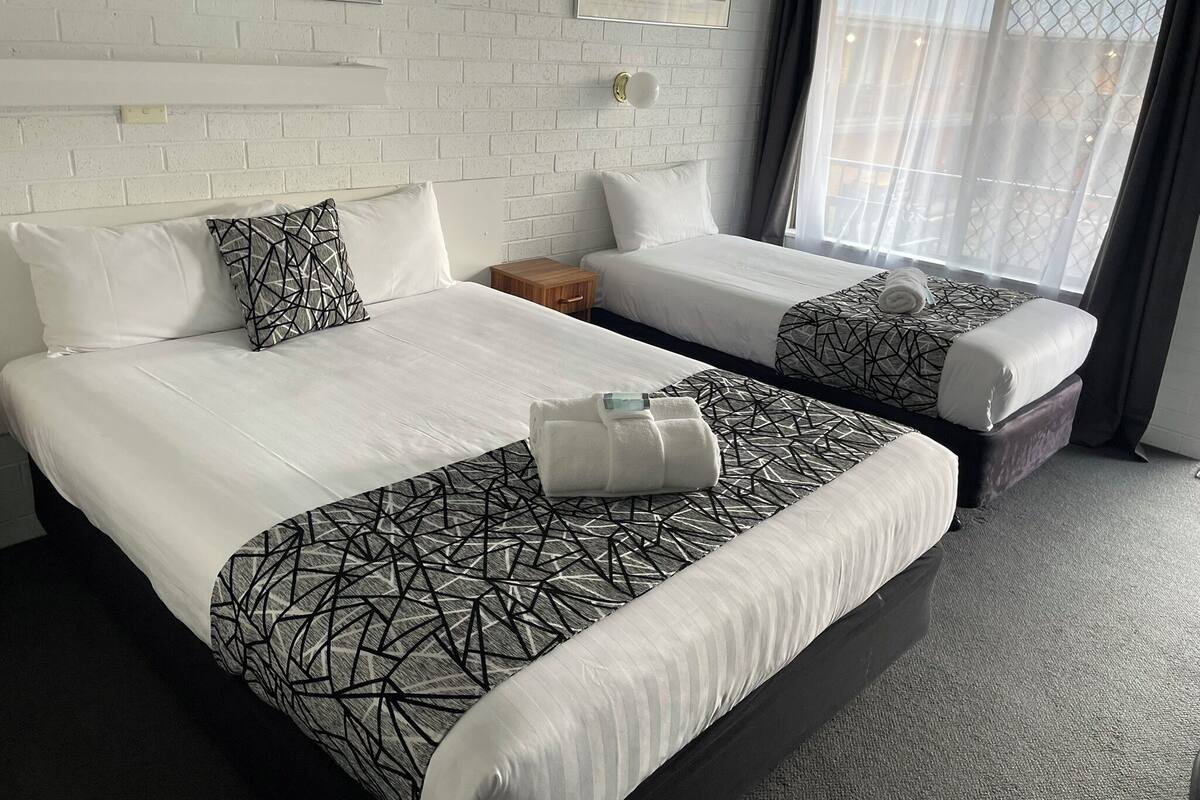 A hotel room with two beds, white bedding, geometric patterned blankets, a small wooden nightstand, and a window with sheer curtains.