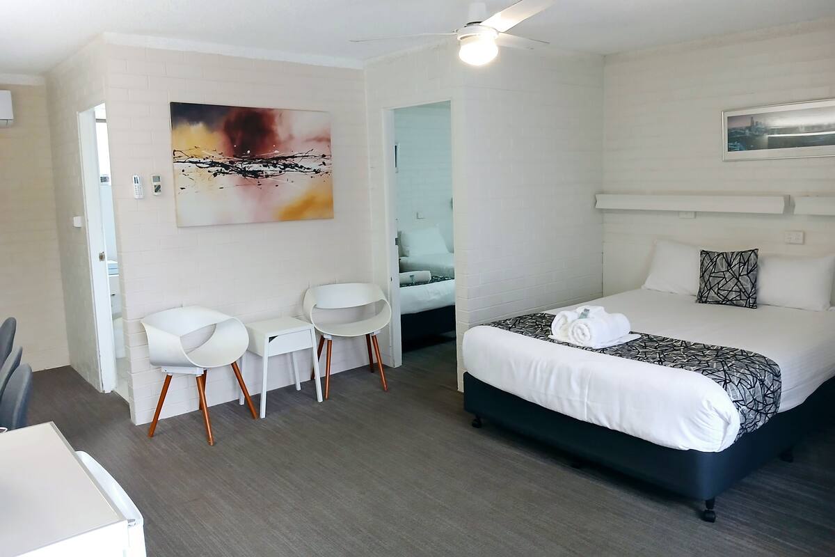 A modern hotel room with a bed, two white chairs, a desk, and a ceiling fan.