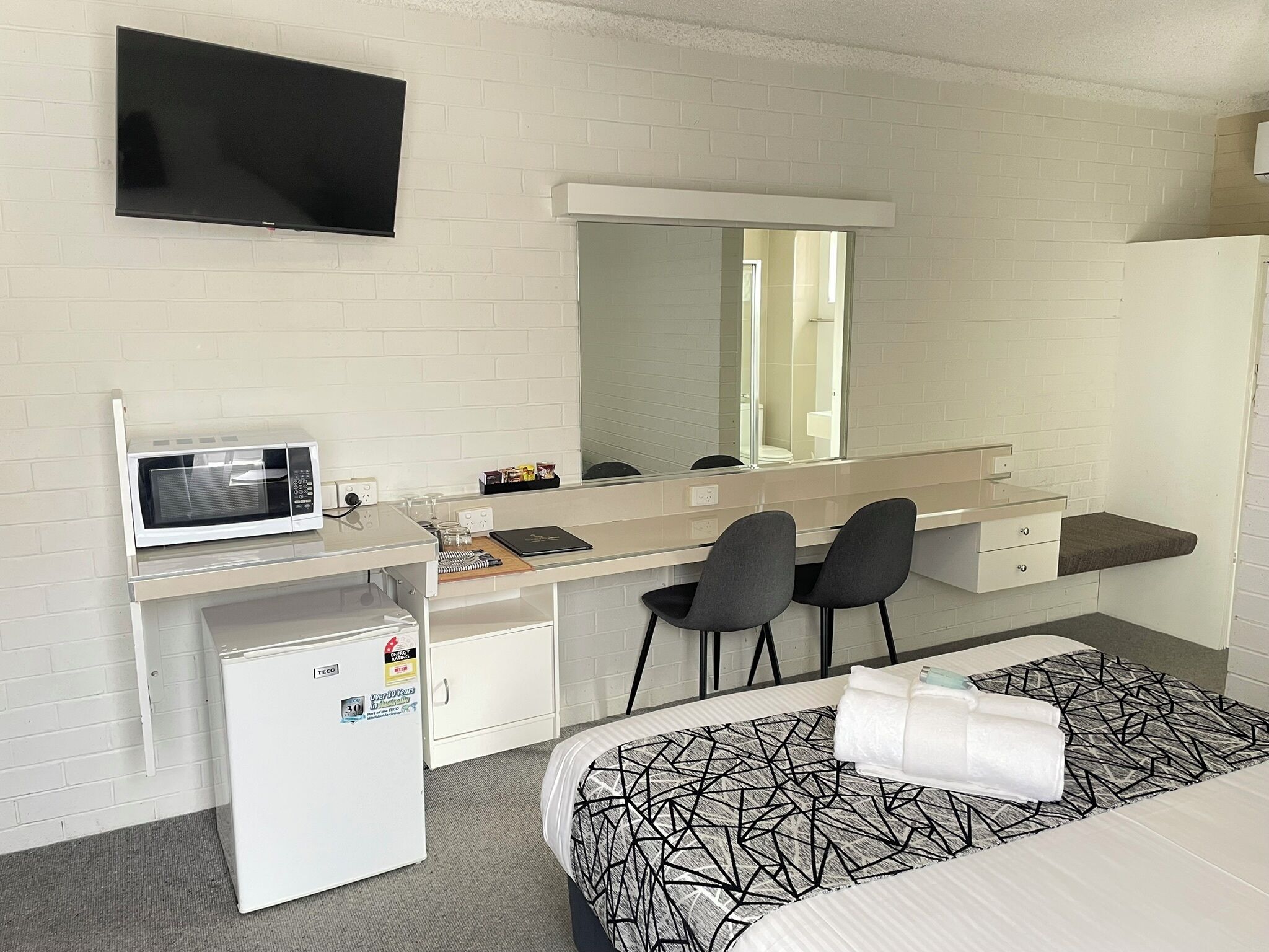 Une chambre d’hôtel équipée d’un lit, d’un bureau avec un micro-ondes, d’un réfrigérateur, d’un miroir et d’une télévision.