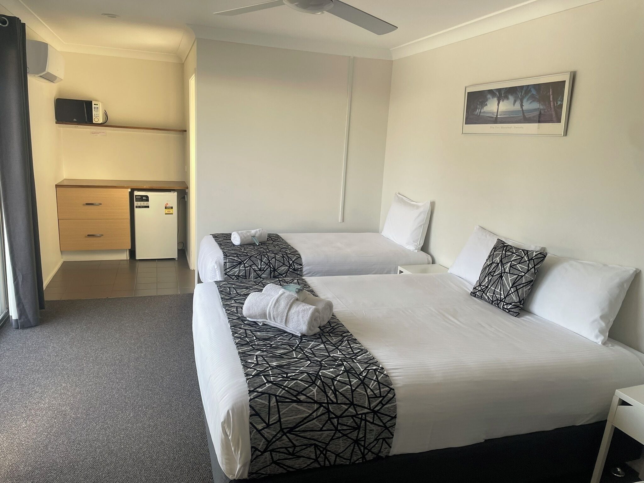 A hotel room with two beds, a small kitchenette, a ceiling fan, and a framed picture on the wall.
