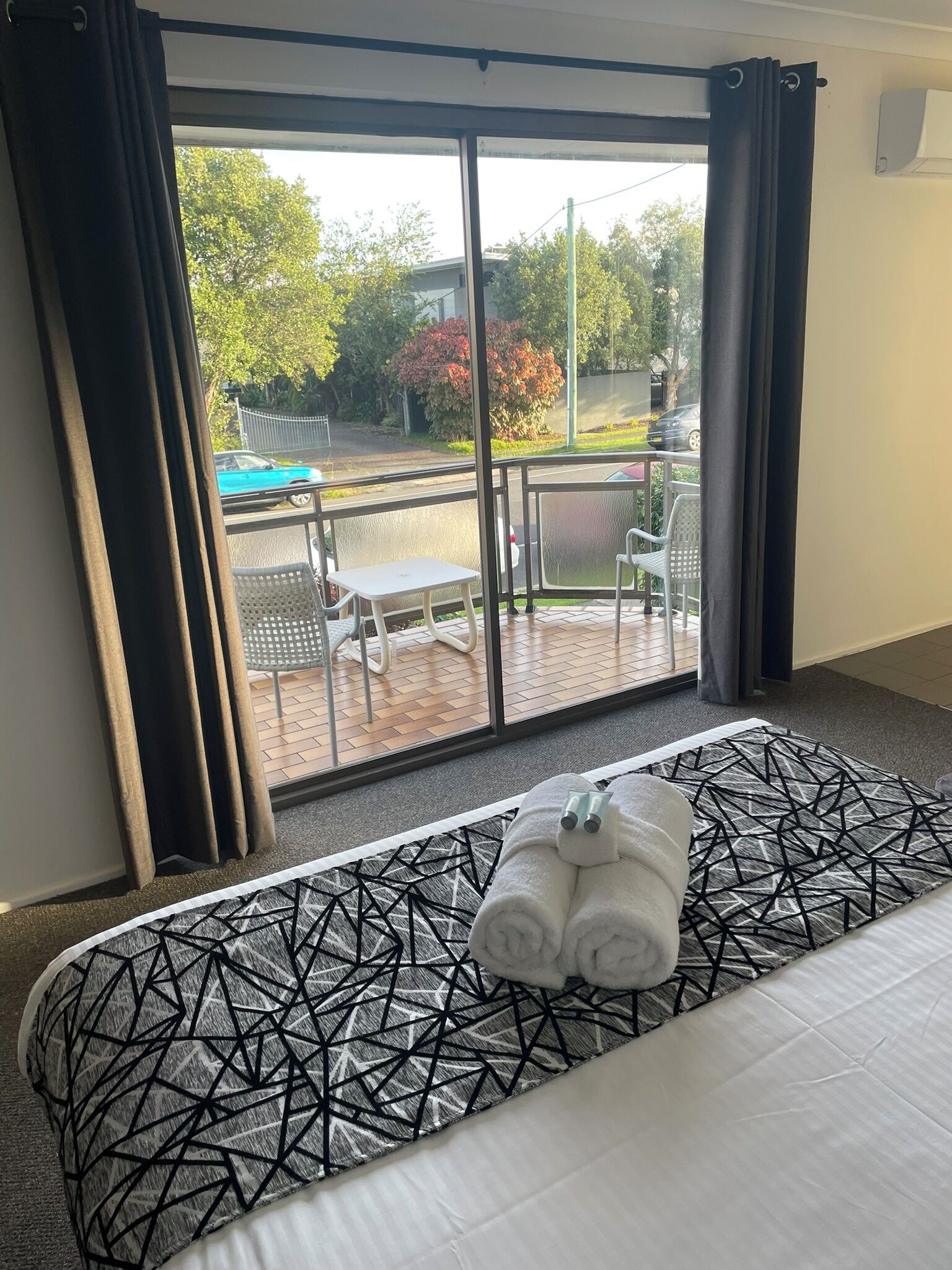 A hotel room with a bed, a view of the outdoor area, and a balcony with chairs and a table.