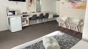 Una habitación de hotel moderna con una cama, un escritorio con un ordenador, un microondas y un refrigerador. Hay dos sillas y un espejo grande sobre el escritorio.