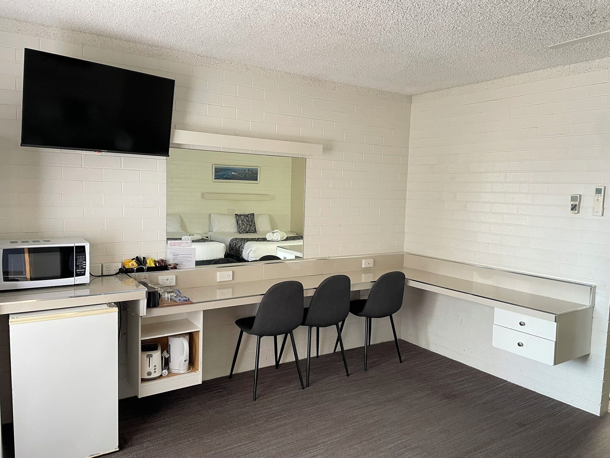 A compact hotel room with a kitchenette, a bed with white bedding, a flat-screen TV mounted above the bed, and a large mirror reflecting the room.