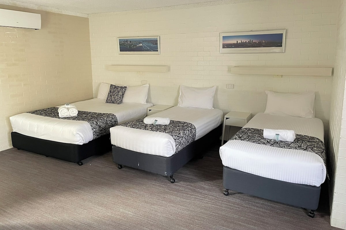 A hotel room with three beds, each with a black and white patterned bedspread, white pillows, and neatly folded towels. There are two framed pictures on the wall, a ceiling fan, and an air conditioning unit on the wall.