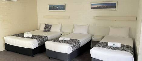 Quarto de hotel com três camas, cada uma com colcha estampada em preto e branco, almofadas brancas e toalhas bem dobradas. Há duas quadros nas paredes, um ventilador de teto e um aparelho de ar condicionado.