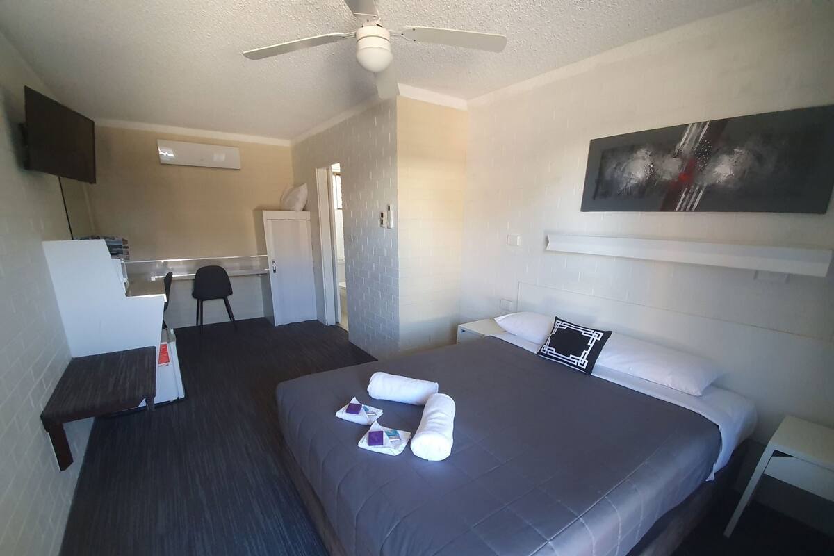 A hotel room with a bed, a desk, a chair, a ceiling fan, and a painting on the wall.