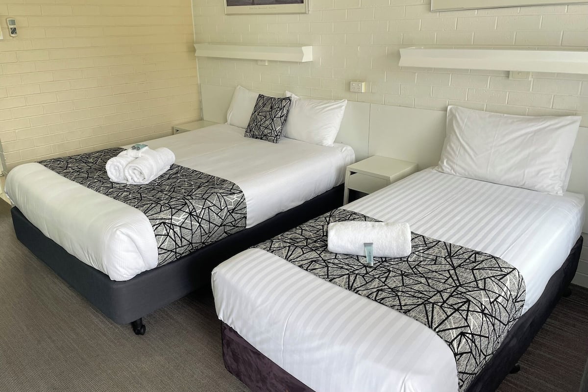 A hotel room with two beds, each with a geometric-patterned bedspread and white pillows. There are framed pictures on the wall, a small bedside table, and a white brick wall.