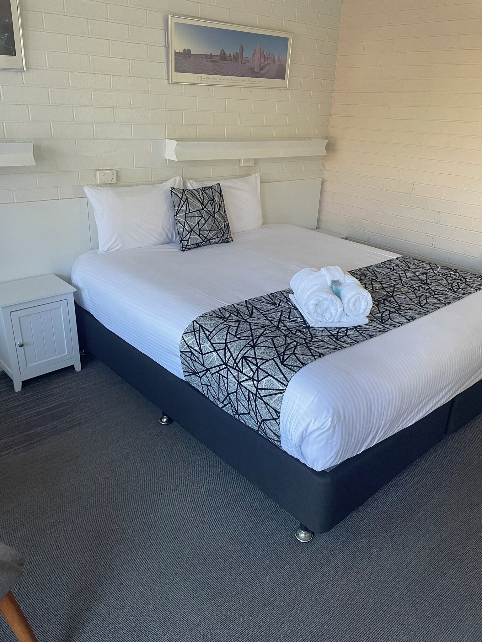A neatly made bed with white linens and a geometric-patterned throw, a small bedside table, and a framed picture on the wall.