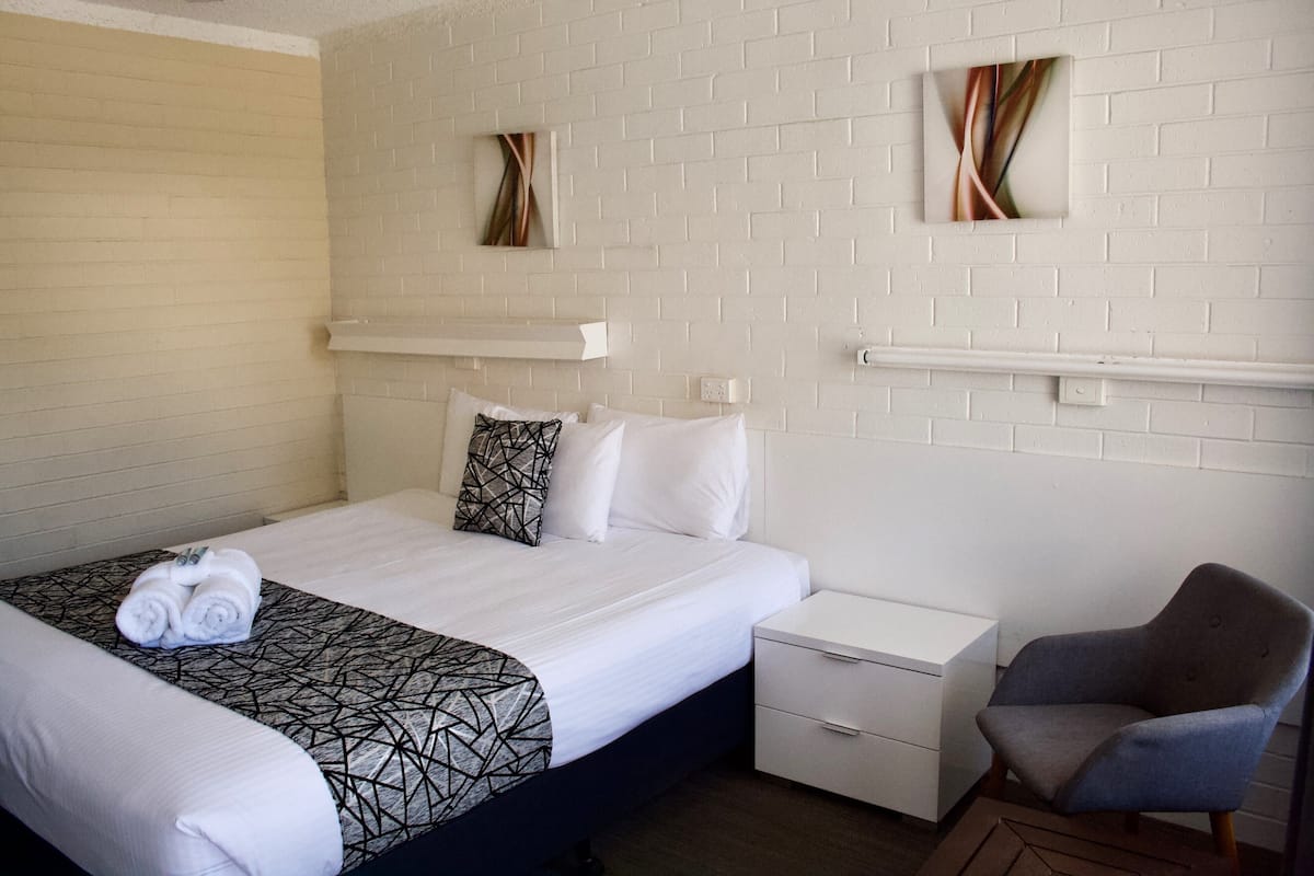 A hotel room with a bed, a chair, a nightstand, and a white brick wall.